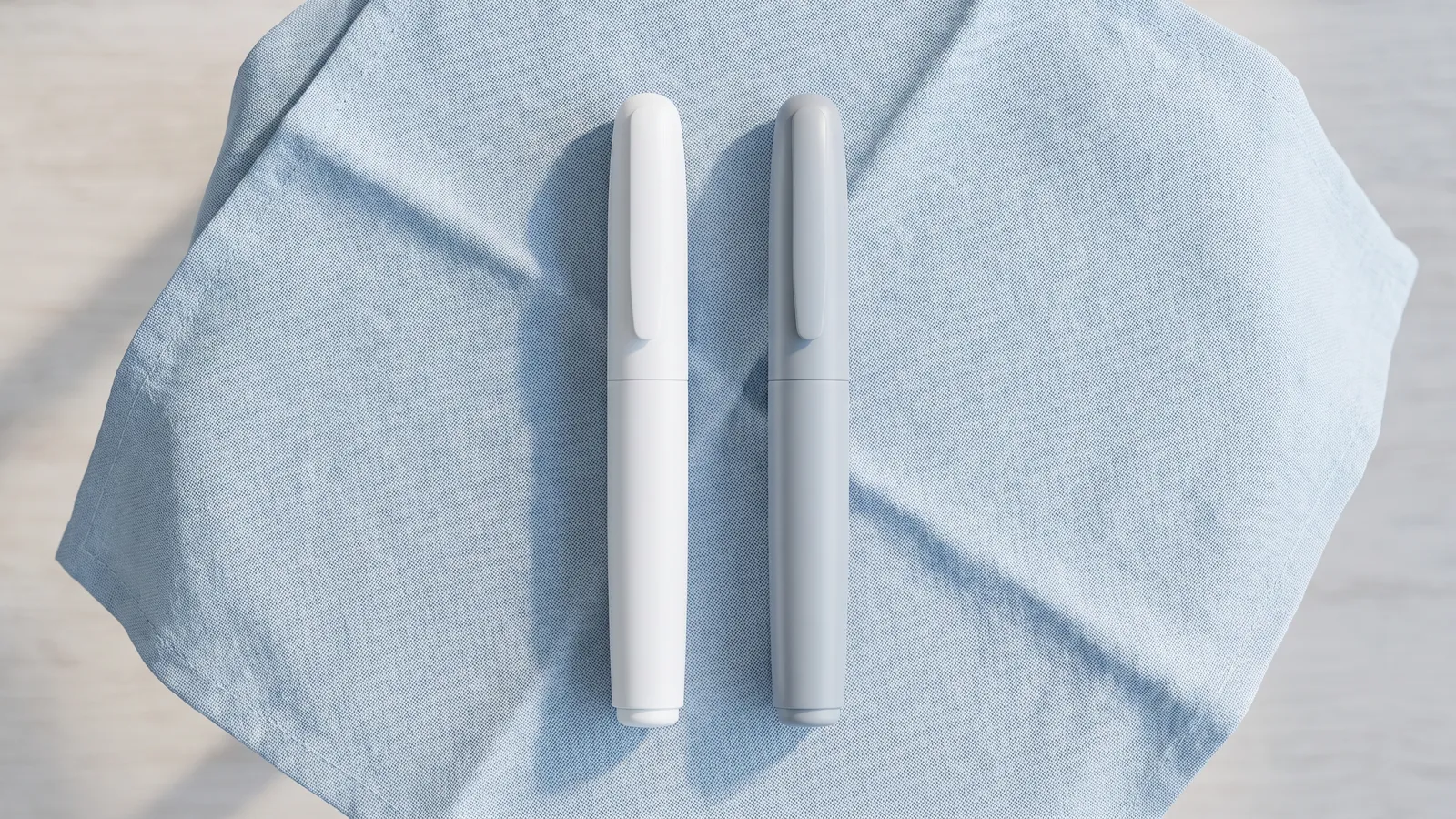 Duas canetas de aplicação sem marca, uma branca e uma cinza-clara, sobre tecido de linho