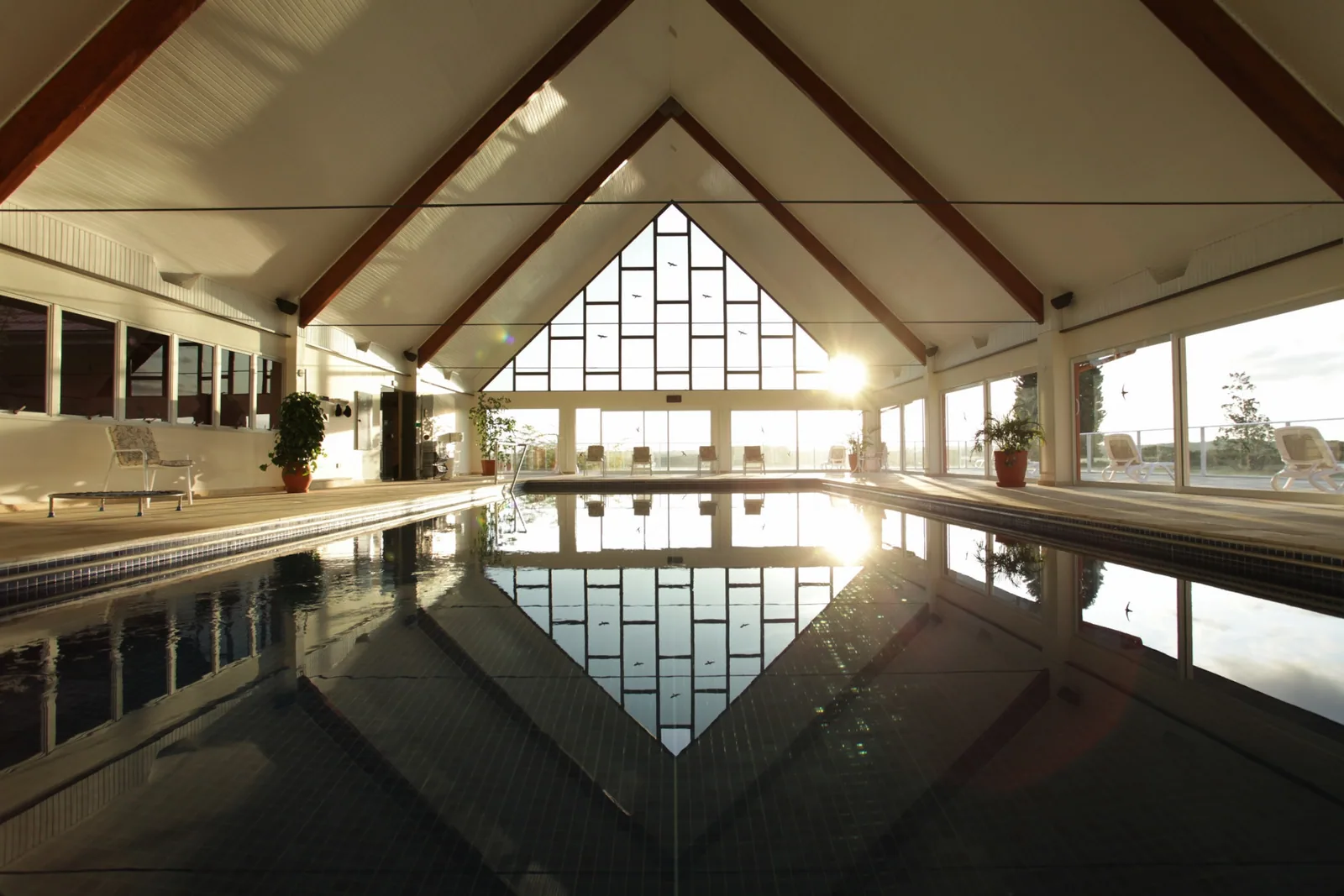 Piscina coberta sob teto de madeira em duas águas, com vitral ao fundo